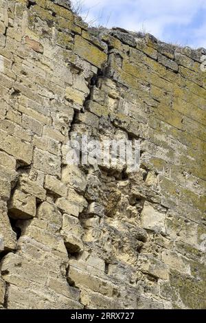 Eine alte, mit Moos bedeckte Steinmauer mit tiefen Rissen und eingestürzten Steinen. Große diagonale Risse an der alten Wand. Leerzeichen kopieren. Selektiver Fokus. Stockfoto