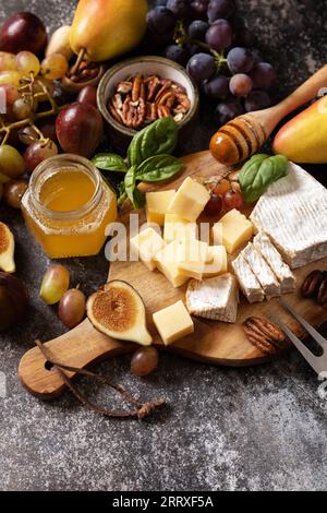 Käseplatte, Stillleben von saisonalen Weinsnacks. Brie-Käse, frische Feigen, Trauben, Käsewürfel, Birnen, Pekannüsse und Honig auf dem Schneidebrett im Dunkeln Stockfoto