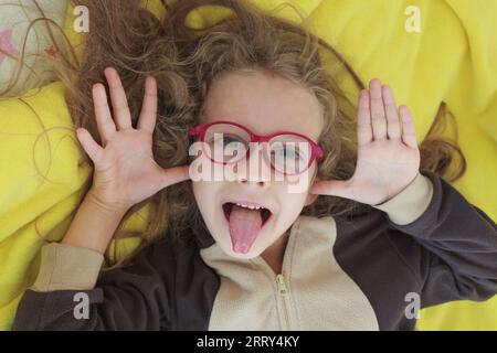 Ein fröhliches Mädchen in der Brille macht Gesichter, neckt und zeigt ihre Zunge mit ihren Händen in Form von Ohren, Nahaufnahme. Stockfoto