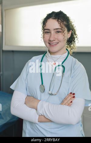 Lächelnde junge Ärztin medizinische Krankenschwester trägt weißen Mantel und Stethoskop um den Hals stehend in der Privatklinik mit den Armen überkreuzt Blick auf Camer Stockfoto