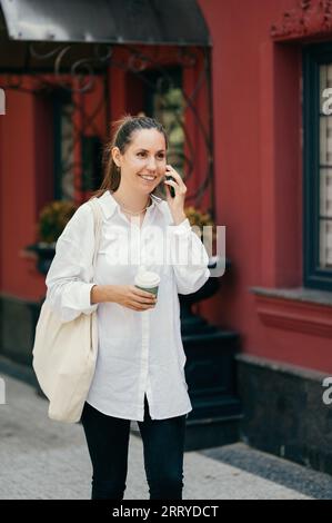 Lächelnde Frau spricht über das Telefon, während sie einen Kaffee und eine Tragetasche hält. Rotes Haus im Hintergrund. Stockfoto