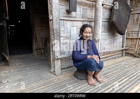 Die einheimische Frau aus dem Stamm der Apatani sitzt auf einem kleinen Stuhl vor einem traditionellen Holzhaus in einem kleinen Dorf in der Nähe von Ziro in Arunachal Pradesh, Indien Stockfoto