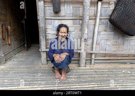 Die einheimische Frau aus dem Stamm der Apatani sitzt auf einem kleinen Stuhl vor einem traditionellen Holzhaus in einem kleinen Dorf in der Nähe von Ziro in Arunachal Pradesh, Indien Stockfoto