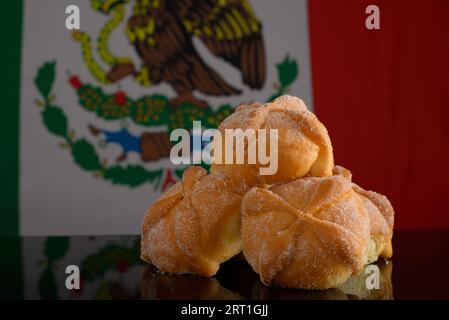 Totes Brot auf schwarzem Glastisch mit mexikanischer Flagge im Hintergrund. Typisches Dessert des Tages der Toten. Stockfoto