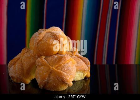 Totes Brot auf schwarzem Glastisch mit farbenfroher Serape im Hintergrund. Typisches Dessert des Tages der Toten. Stockfoto