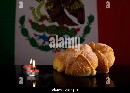 Totes Brot und Kerzen auf schwarzem Glastisch mit mexikanischer Flagge im Hintergrund. Typisches Dessert des Tages der Toten. Stockfoto