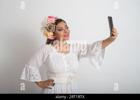 Mexikanische Frau mit Blumenkopfbehaarung im Haar, die ein Selfie macht. Weißer Hintergrund. Stockfoto