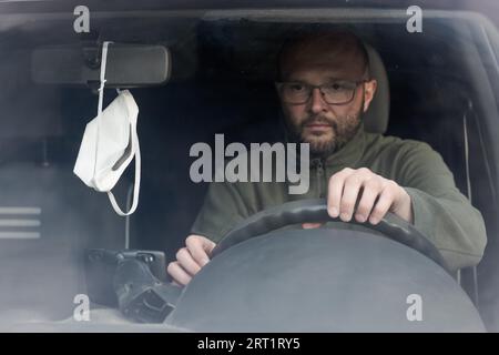 Porträt eines jungen Mannes, der im Auto sitzt und auf eine Schutzmaske schaut, die vom Rückspiegel im Auto hängt, von vorne gesehen Stockfoto