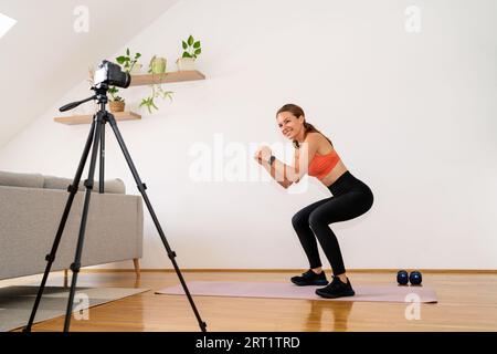 Junge Erwachsene Sportlerin, die zu Hause vor einer Videokamera auf einem Stativ Squats macht. Stockfoto