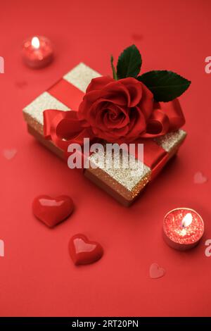 Goldene Geschenkbox mit Band und der Rose auf rotem Hintergrund, Valentinskarte oder Muttertagskarte Kopierraum flach liegend, vertikal Stockfoto