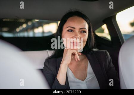 Lächelnde, hübsche Geschäftsfrau sitzt auf dem Rücksitz eines Taxis und hält die Hand am Kinn. Geschäftskunden. Konzept für Autovermietung Stockfoto