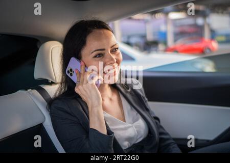 Glückliche Geschäftsfrau, die auf der Rückbank eines Autos sitzt, während sie mit dem Smartphone telefoniert. Business, Roadtrip, Technologie, Reisekonzept Stockfoto