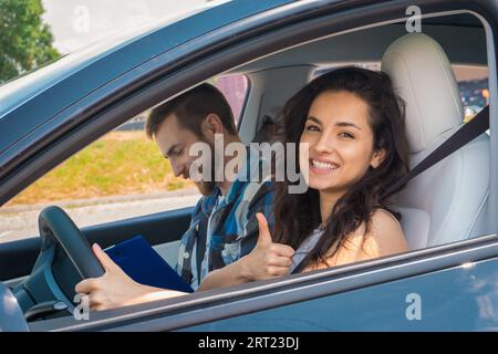 Lächelnder männlicher Ausbilder, der die Prüfung bei der jungen Dame ablegt. Glückliche Frau sitzt im Auto und zeigt Daumen hoch. Fahrprüfung, Fahrerkurse, Prüfungskonzept Stockfoto