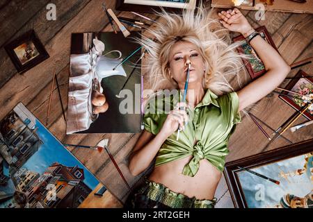 Blick von oben auf eine junge blonde Malerin, umgeben von ihren Kunstwerken Stockfoto