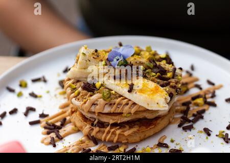 Ein Stapel Bananenpfannkuchen mit Scheiben frischer Bananen, Pistazien und Honig, dazu Schokoladenscheiben und essbare Blume. Nahaufnahme Stockfoto