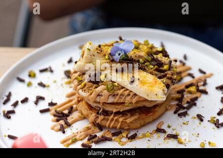 Ein Stapel Bananenpfannkuchen mit Scheiben frischer Bananen, Pistazien und Honig, dazu Schokoladenscheiben und essbare Blume. Nahaufnahme Stockfoto