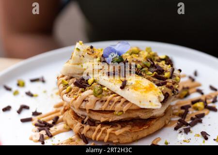 Ein Stapel Bananenpfannkuchen mit Scheiben frischer Bananen, Pistazien und Honig, dazu Schokoladenscheiben und essbare Blume. Nahaufnahme Stockfoto