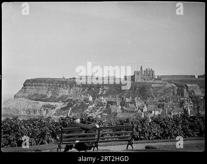 Whitby, Scarborough, North Yorkshire, 1925-1935. Ein Blick auf Whitby Abbey von West Cliff, mit einem Paar, das auf einer Bank im Vordergrund sitzt. Stockfoto