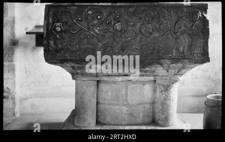 All Saints Church, East Meon, East Hampshire, Hampshire, 1940-1949. Detail einer Tournai-Schriftart mit einer quadratischen Schüssel mit Schnitzereien von Figuren in der Allerheiligen-Kirche. Das Bild zeigt eine ganze Seite der Tournai-Schriftschale mit Figuren und einigen Tiergesichtern mit flachen Markierungen. Tournai-Schriftarten sind aus einem schwarzen Stein geschnitzt, der in Tournai, Flandern, gefunden wurde. Die Schnitzereien zeigen die Geschichte von Adam und Eva, von Adams Schöpfung und der Schöpfung von Eva von Adams Rippe auf der rechten Seite bis hin zu Eva, die Früchte von einer Schlange auf der linken Seite nimmt. Die Schüssel sitzt auf einem kreisförmigen Ziegelschacht, der dicht von vier Säulen umgeben ist. Stockfoto