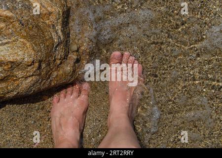 Barfuß-Frauenfüße aus nächster Nähe im klaren Wasser auf dem Sandboden neben einem Felsen. Stockfoto