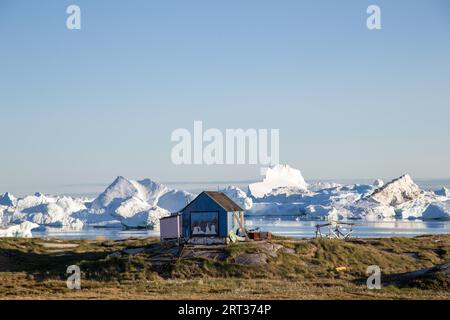 Rodebay, Grönland, 9. Juli 2018: Ein blaues Holzhaus mit Eisbergen im Hintergrund. Rodebay, auch bekannt als Oqaatsut, ist eine Fischersiedlung im Norden Stockfoto