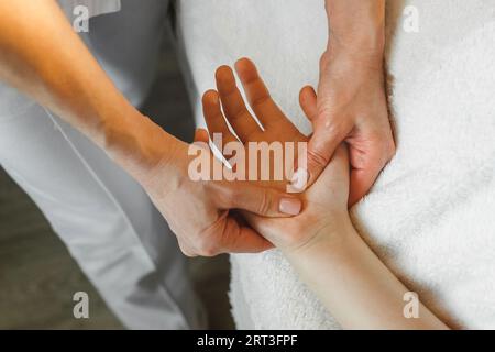 Nahaufnahme einer Masseurin, die Arm, Handgelenk, Finger und Handfläche einer Klientin massiert, die auf einem Massagetisch in einem Salon liegt. Stockfoto