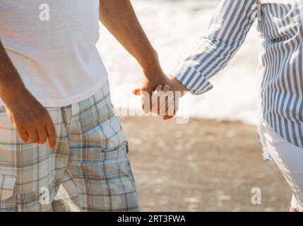 Ein älteres Ehepaar geht an der Küste entlang und ruht sich aus. Meereswellen im Hintergrund. Nahaufnahme der Hände. Stockfoto