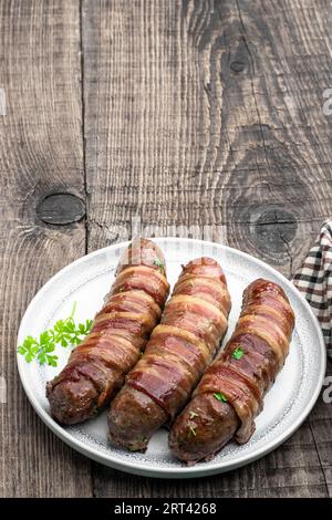 Appetitliche Würstchen in Speck gewickelt und auf weißem Teller auf Holztisch gegrillt Stockfoto