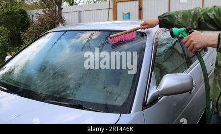 Ein Mann in einer grünen Jacke wäscht ein silbernes Auto aus einem Gummischlauch und reinigt es mit einer rosa Bürste im Innenhof eines Privathauses mit Selbstbedienung Stockfoto