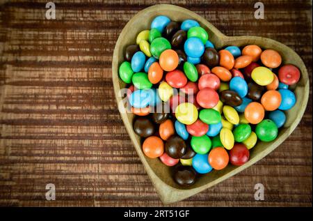 Haufen farbenfroher Schokoladenbonbons in herzförmiger Holzschale auf hölzernem Hintergrund, Draufsicht Stockfoto