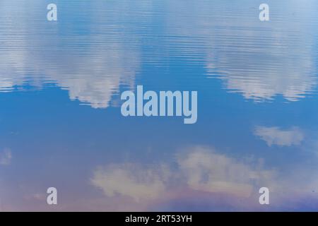 Blauer Himmel und bewölkter, farbenfroher Sonnenuntergang, der sich im stillstehenden Wasser am Strand widerspiegelt. Gras, Bäume, Schilf und Himmelsreflexion im Wasser Stockfoto