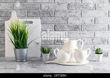 Schöner stilvoller Küchenhintergrund in hellen Farben. Ein Set aus weißen Krügen, Schneidebrettern und Zimmerpflanzen auf einer grauen Steinarbeitsplatte. Graue Ziegelwand Stockfoto