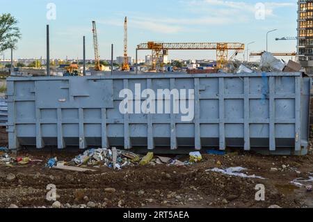 Großer Metallbehälter zur Sammlung von Bauabfällen. Im Hintergrund befindet sich ein Baukran in oranger Farbe. Groß, nicht im Haushalt, Stockfoto