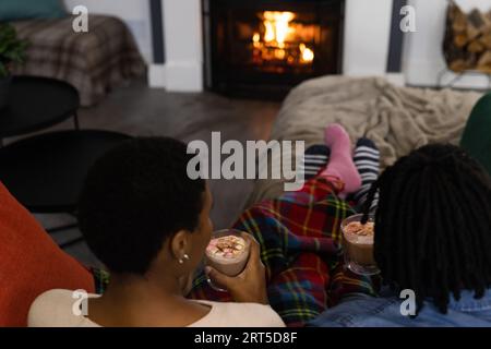 afroamerikanisches Paar in Socken, das sich zu Hause entspannt und vor offenem Feuer heiße Schokolade trinkt Stockfoto