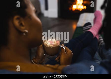 afroamerikanisches Paar in Socken, das sich zu Hause vor dem offenen Feuer mit heißer Schokolade entspannt Stockfoto