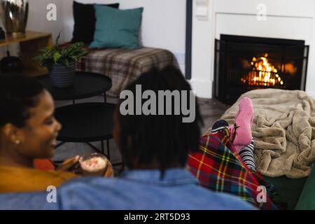 Glückliches afroamerikanisches Paar in Socken, das sich zu Hause vor dem offenen Feuer mit heißer Schokolade entspannt Stockfoto