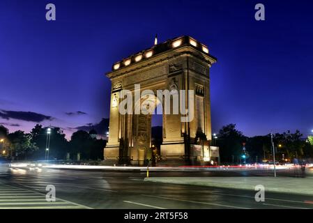 Arcul de Triumf an der Kiseleff Road, Bukarest, Rumänien. Die leichte Spur des abendlichen Pendlerverkehrs ließ einen vollen Kreis am Sonnenuntergang brennen Stockfoto