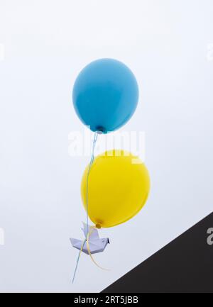 Feuern Sie eine Papiertaube des Friedens ab, die an zwei gelb-blaue Ballons in der Farbe der ukrainischen Flagge gebunden ist. Unterstützung Der Ukraine. Hör auf mit dem Krieg. Symbol des Friedens und f Stockfoto