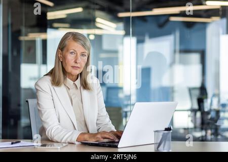 Porträt einer seriösen, reifdenkenden Geschäftsfrau, grauhaariger weiblicher Boss, der in die Kamera schaut, am Arbeitsplatz im Büro, Finanzier mit Laptop, selbstbewusster Buchhalter. Stockfoto