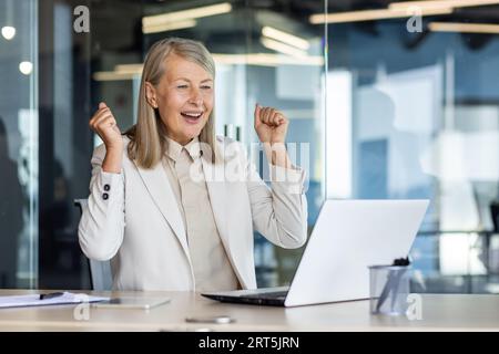 Senior erfolgreiche Geschäftsfrau feiert Siegeserfolg, grauhaarige Arbeitsplatzfrau, die die Hände hoch hält, zufrieden mit den Arbeitsergebnissen, Frau Chefin im Business-Anzug. Stockfoto