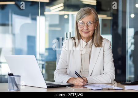 Porträt einer älteren, Reifen Finanzierin am Arbeitsplatz, grauhaarige Geschäftsfrau, die lächelt und die Kamera anschaut, die Papierkram macht, Boss im Business-Anzug mit Laptop im Büro. Stockfoto