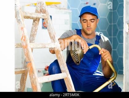 Ein erfahrener Arbeiter mit blauen Overalls sitzt in der Hocke und repariert den Duschkopf im Badezimmer Stockfoto