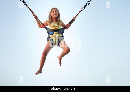 Bungee-Jumping auf Trampolin. Kleines Mädchen hüpft auf Bungee-Jumping im Vergnügungspark auf Sommerferien. Kind fliegt über den blauen Himmel, während es weiterspringt Stockfoto