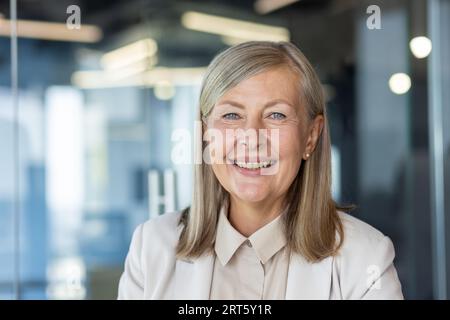 Nahaufnahme einer Reifen grauhaarigen Geschäftsfrau, einer älteren erwachsenen Frau, die lächelt und die Kamera anschaut, am Arbeitsplatz im Büro. Stockfoto