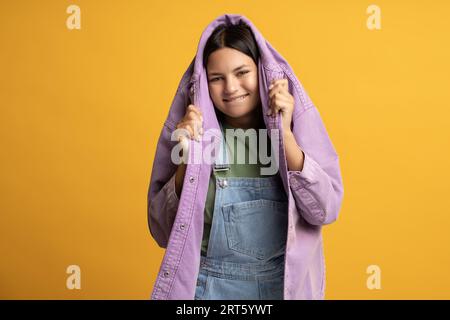 Glückliches lächelndes, verspieltes Teenager-Mädchen, das sich mit Kopf in einer lila Jeansjacke auf gelbem Hintergrund versteckt. Stockfoto