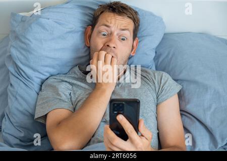 Nervöser, beeindruckter Mann, der Nachrichten auf dem Smartphone las und morgens im Bett einen besorgten Ausdruck hatte. Guy staunte über schlechte Nachrichten, die auf dem Handy empfangen wurden Stockfoto