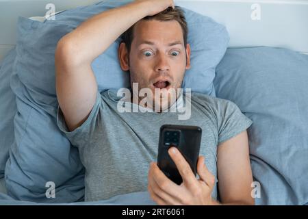 Gut aussehender beeindruckter Mann, der am Morgen Nachrichten auf dem Smartphone las, mit schockiertem Ausdruck im Bett. Guy staunte über die Nachrichten, die auf dem Handy empfangen wurden, Stockfoto