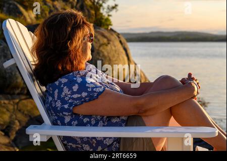 Eine Frau mittleren Alters, die Indianer anständig ist, sitzt auf einem Adirondack-Holzstuhl auf einem Steg, der der untergehenden Sonne im Kragero-Archipel, Telemark, Norwegen, zugewandt ist. Stockfoto