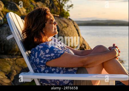 Eine Frau mittleren Alters, die Indianer anständig ist, sitzt auf einem Adirondack-Holzstuhl auf einem Steg, der der untergehenden Sonne im Kragero-Archipel, Telemark, Norwegen, zugewandt ist. Stockfoto