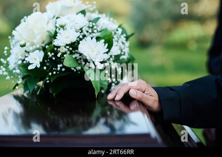Konzentrieren Sie sich auf die Hand einer Trauernden Reifen Frau in schwarzer Kleidung auf dem Deckel eines geschlossenen Holzsarges mit einer Menge frischer weißer Chrysanthemen auf der Oberseite Stockfoto
